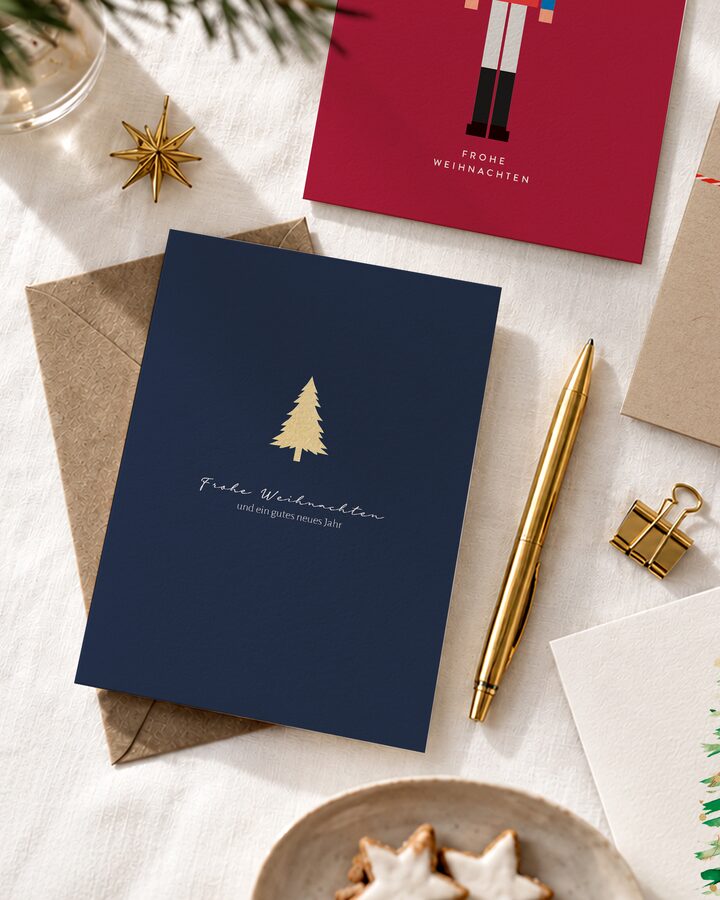 Dunkelblaue Weihnachtskarte mit goldener Tanne auf Kraftpapier-Umschlag, daneben goldener Stift, weitere Karten und Tannenzweig auf weißem Leinen